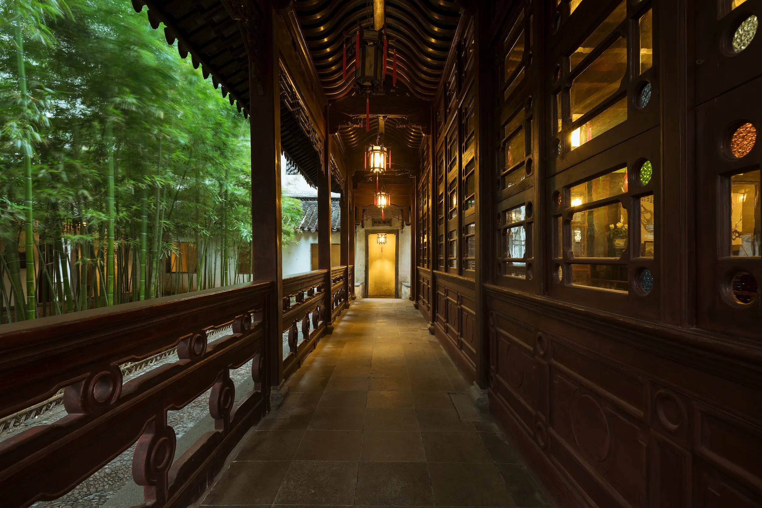 Covered wooden corridor with carved details and glowing lanterns, beside a swaying bamboo grove, in Lion Grove Garden, Suzhou.