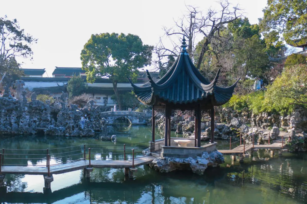 Traditional Chinese pavilion with a multi-eaved roof surrounded by rockeries and a pond in Lion Grove Garden, Suzhou. A winding stone bridge and lush greenery complete the tranquil landscape.