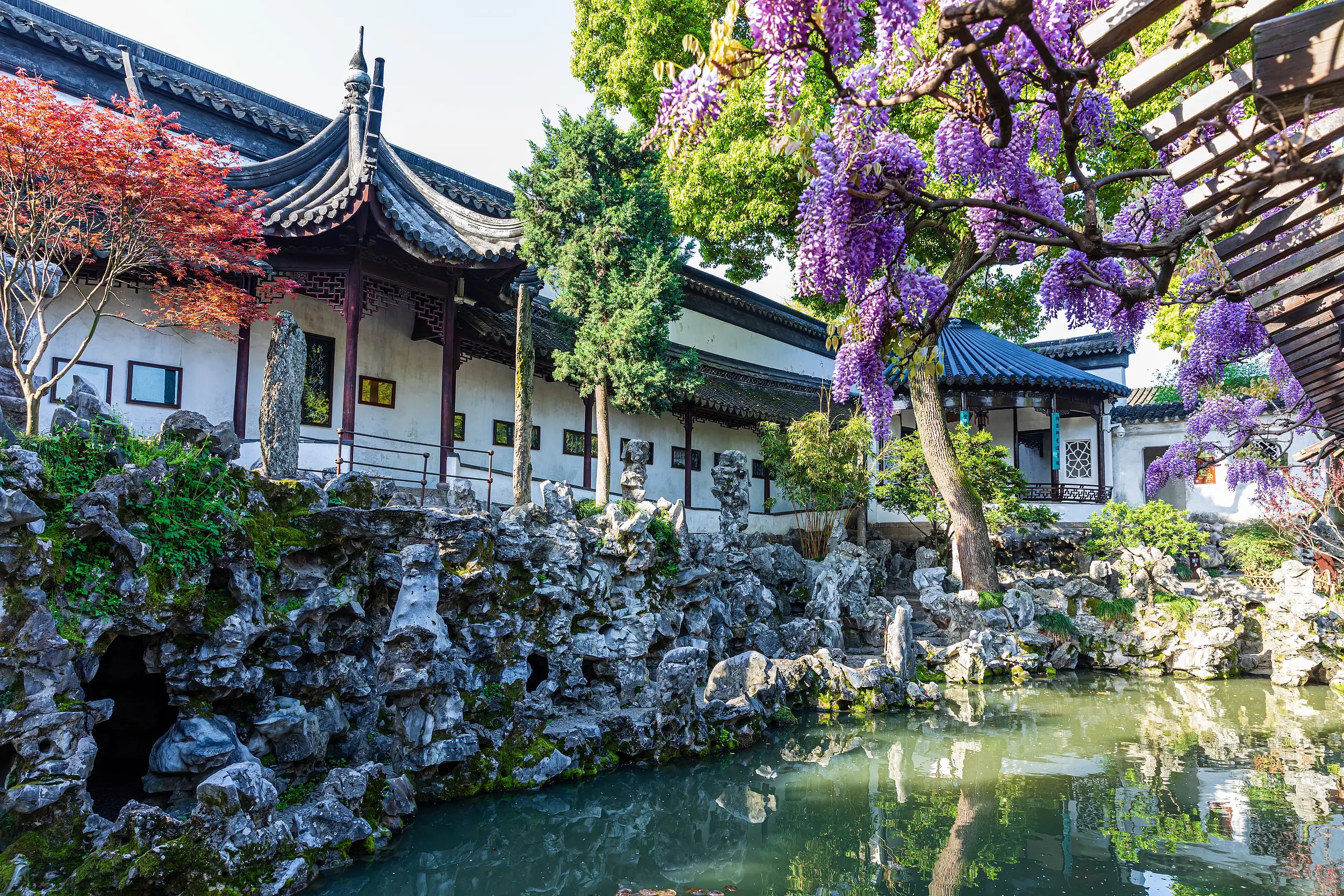 Scenic view of Lion Grove Garden in Suzhou with traditional Chinese architecture, blooming wisteria, unique rockeries, and a still pond reflecting the serene landscape.