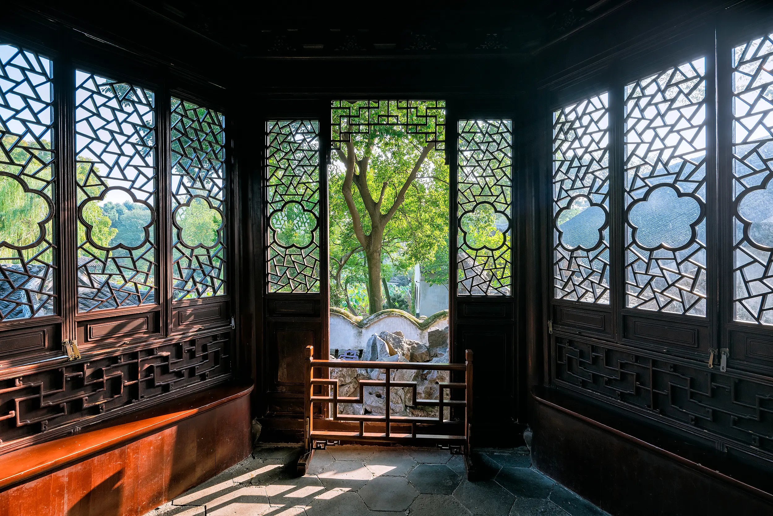A classical Chinese room in the Humble Administrator’s Garden (Zhuozhengyuan), Suzhou. The interior features traditional wooden furniture and a stone-tiled floor in hexagonal patterns. Intricately designed lattice windows filter soft daylight while revealing glimpses of a lush green courtyard outside. The space exudes a tranquil and scholarly ambiance, evoking the refined lifestyle of ancient literati.