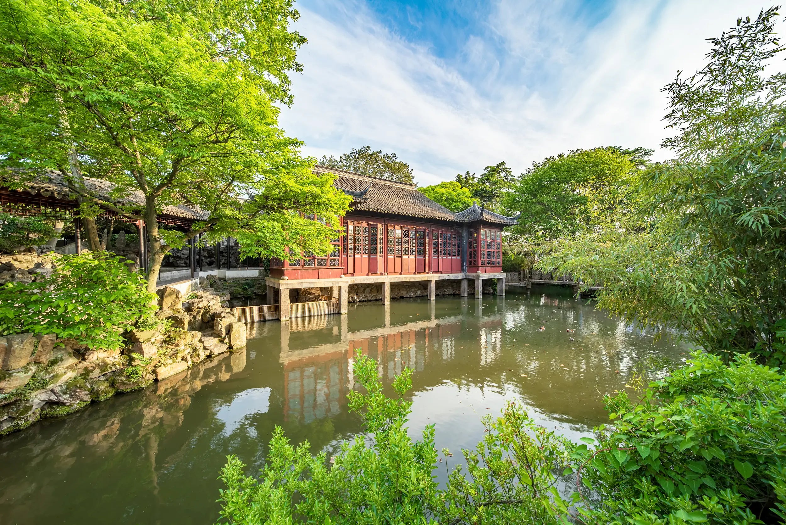 A red pavilion stands gracefully above a tranquil pond, surrounded by lush greenery. The still water reflects the vibrant architecture and the blue sky with white clouds, creating a serene and harmonious scene in Suzhou’s classical garden.