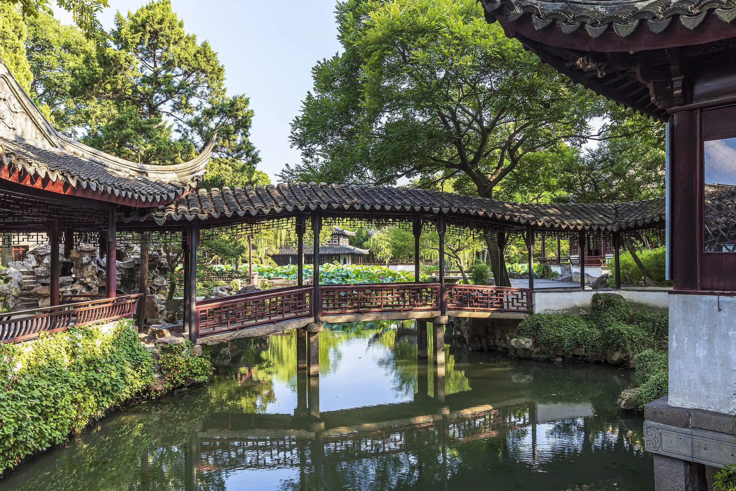 A winding zigzag corridor stretches above a clear pond, connecting pavilions and scenic spots through elegant bridges. Surrounded by lush greenery and traditional Chinese architecture, the scene embodies the tranquil charm and intricate design of Suzhou’s classical garden. Reflections on the water enhance the natural harmony and artistic depth.